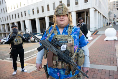 Mies seisoo aukiolla rynnäkkökiväärin kanssa. Hänellä on päällään Havaiji-paita sekä armeijan taisteluliivi.