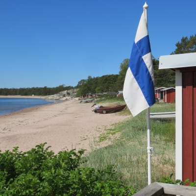 En solig sommardag vid Gunnarsstrand i Hangö. Sandstrand, knallblå himmel, miniflagga (Finlands) vid en fiskebod. Flera fiskebodar och båtar längs stranden.