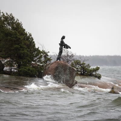 naista esittävä patsas kalliolla meren rannalla, myrsky lyö aaltoja kallioon