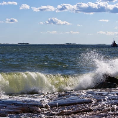Vågor som slår mot en klippig strand på Sveaborg. Vid horisonten syns en segelbåt.
