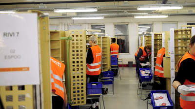 Postens anställda iklädda orange västar står i bås och sorterar post.
