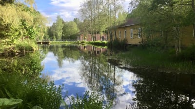 Gamla slitna industribyggnader vid vatten i ett brukssamhälle. Sommar, solsken, grönt gräs och gröna träd. Byggnaderna och den lummiga naturen reflekteras i vattnet.