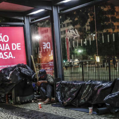 En hemlös man sover vid en busshållptas intill en skylt med texten "Gå inte ut från ditt hem"
