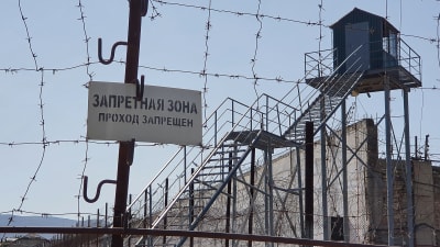 I förgrunden syns taggtråd och stängsel samt en skylt på ryska. Bakom stängslet en trappa i stål som leder upp till ett vakttorn.