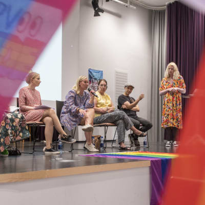 Sex personer deltar i en paneldiskussion på en scen. En av dem pratar i en mikrofon. I förgrunden syns regnbågsflaggor.