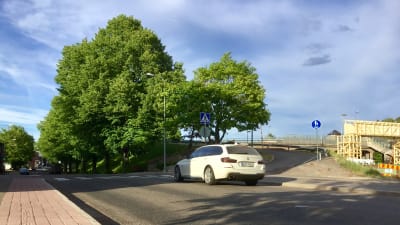 En gata med lövträd vid vägrenen invid en brobank. En vit bil kör på gatan. Bron syns till höger. Sommar,