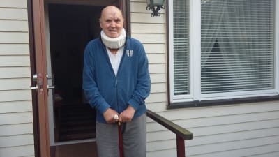 En äldre man med nackstöd och sår i pannan står på en trappa utanför ett vitt hus i trä.
