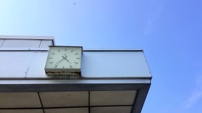 En analog väggklocka på fasaden av en sliten vit marmorbyggnad. Det är antagligen en 1970-talsklocka som finns kvar på busstationens vägg i Karis.