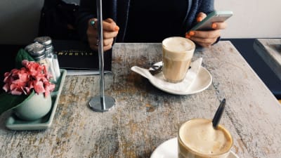 Två koppar kaffe på bordet i ett kafée och en kvinnohand som håller i en mobiltelefon.