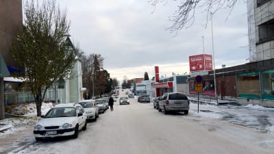 En snöbeklädd väg. På sidorna syns parkerade bilar och till höger syns en butik.