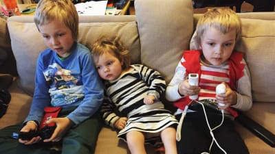 Johannes Romppanens barn Emil och Caper spelar konsolspel i soffan och lillasyster Lilja sitter i mitten och följer med.