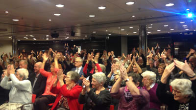 en stor samling pensionärer klappar i händerna i stor sal med röda sitsar 