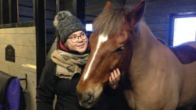 En kvinna står bredvid en häst i ett stall. Hon håller handen runt hästens mule.