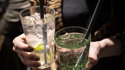 Vasemmalla käsi pitelee väritöntä drinnkiä, jossa on sitruuna- tai limelohko. Oikealla on kädessä vihreä drinkki. Molemmissa laseissa on pilli. 