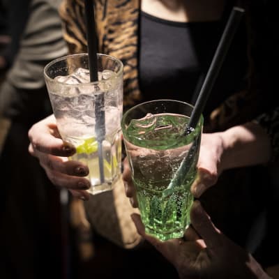 Vasemmalla käsi pitelee väritöntä drinnkiä, jossa on sitruuna- tai limelohko. Oikealla on kädessä vihreä drinkki. Molemmissa laseissa on pilli. 