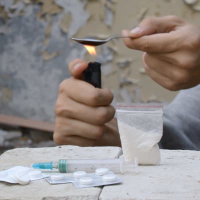 En person sitter och värmer något preparat i en sked. På bordet framför syns en påse med vitt pulver, en spruta och några ark med tabletter.