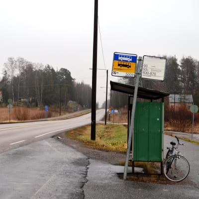 En busshålplats intill en landsväg sedd från sidan. Inga människor syns i bild, en cykel står parkerad bakom busskuren.