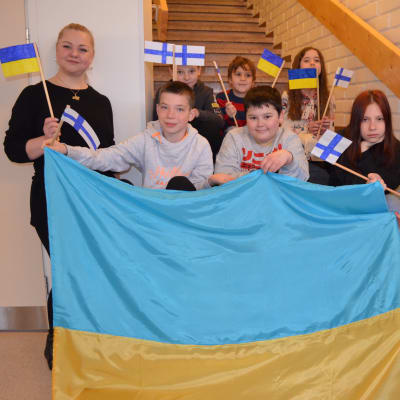 Joukko lapsia ja kaksi naista pitävät kiinni Ukrainan lipusta ja heiluttavat samalla käsissään pieniä Ukrainan ja Suomen lippuja.