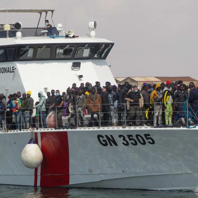 Kymmeniä afrikkalaisia siirtolaisia laivan kannella saapumassa satamaan. Laivan kyljessä lukee "Garde Nationale".