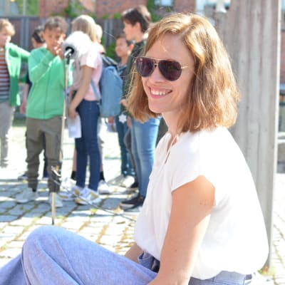 Redaktör Sandra Strömvall sitter leende med solglasögon på skolgård med skolbarn i bakgrunden.