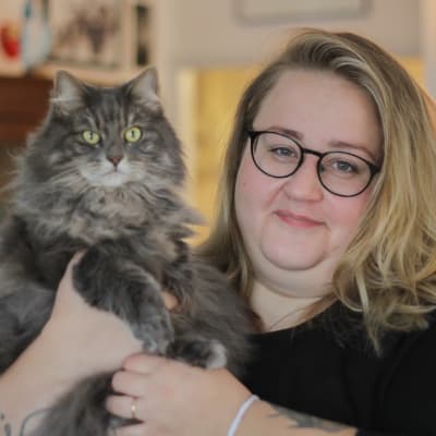En vit kvinna med blont hår och svarta glasögon ler och håller upp en långhårig, grå katt.