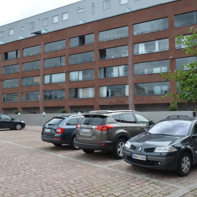 Ett höghus i sex våningar i rödbrunt tegel sett framifrån. Framför i bilden syns parkeringsrutor varav ungefär hälften är upptagna av vanliga personbilar.