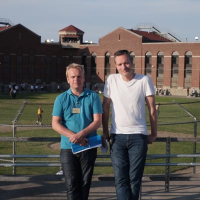 Två män står framför en tegelbyggnad med en sportplan i bakgrunden och metallstaket runt. 