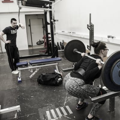 En kvinna och en man i ett gym. Kvinnan gör knäböj med en stång och vikter på axlarna som väger sammanlagt 80 kg. Mannen tittar på.