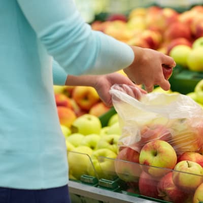 Person plockar frukter i plastpåse. I bakgrunden syns flera lådor av äpplen. 