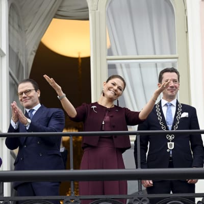 Kronprinsessan Victoria och prins Daniel på Rådhusets balkong i Lovisa tillsammans tre andra personer, en kvinna och två män. Daniel applåderar och Victoria sträcker ut armarna i uppskattning av alla de människor som samlats på torget nedanför stadshuset.