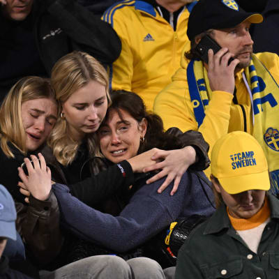 Tre upprörda kvinnor kramar om varandra på en läktare bland gulklädda svenska fotbollsfans.