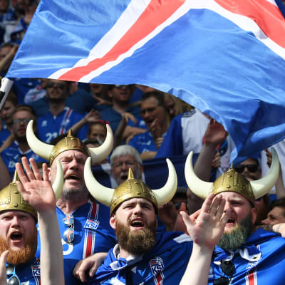 Islantilannin lipun väreihin pukeutuneita jalkapallofaneja. Etuan miesjoukolla on viikinkihatut ja he heiluttavat Islannin lippua.