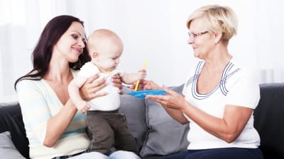en ung kvinna sitter i en soffa, har ett litet barn i famnen, en äldre dam sitter bredvid henne och ger en leksak till barnet. 
