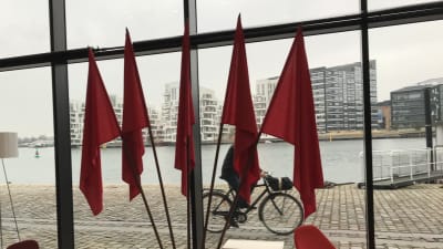 Fem röda flaggor framför ett stort fönster med utsikt över Köpenhamn.