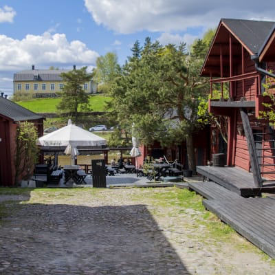 En sommarrestaurang mitt emellan två gamla hus. I bakgrunden skymtar en å och ett stort gult trähus på en kulle.