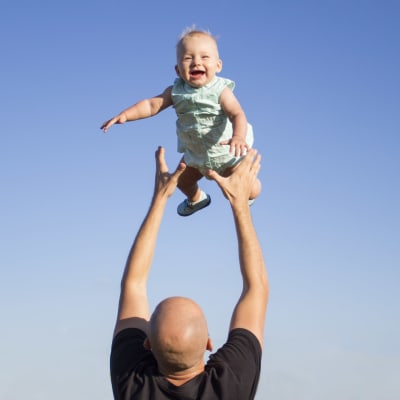 Isä leikittää pientä lasta heittämällä häntä korkealle ilmaan. Lapsi nauraa iloisena. Lapsen taustalla näkyy sininen pilvetön taivas.