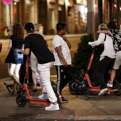 En person åker elsparkcykel på trottoar på natten. Två personer bakom honom åker på samma elsparkcykel. 