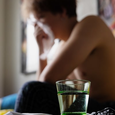 Längst fram på bilden ett vattenglas och värktabletter, i bakgrunden en anonymiserad ung man med bar  överkropp som sitter på en säng och snyter sig.