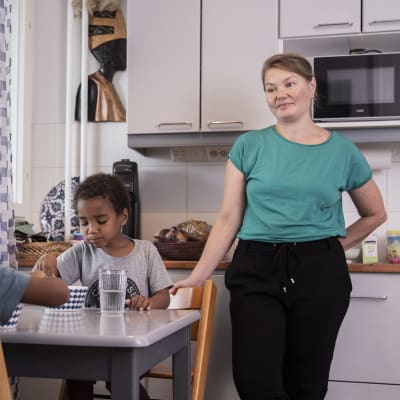 En mamma står bredvid ett köksbord, ser på sina två barn som äter. 