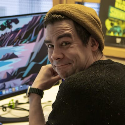Sami Saari istuu työpöytänsä ja -koneensa äärellä ja katsoo olkansa yli kameraa kohti. 