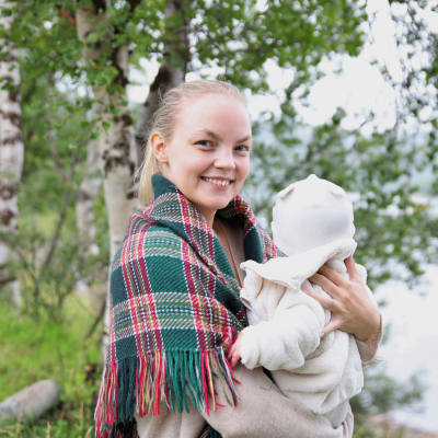 Miisa Nuorgam katsoo kameraan vauva sylissään, taustalla näkyy lehtevien koivunoksien läpi jokimaisema.