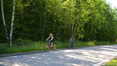 Kaarina Hurtta och Sari Siikala cyklar längs med Eriksnäsvägen. I bakgrunden grön skog.