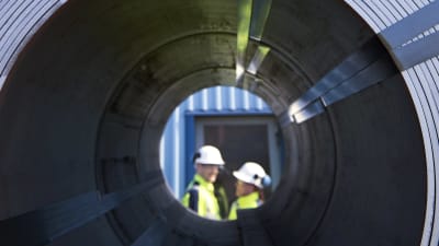 Stålrulle och två personer i reflexkläder och vita hjälmar syns på andra sidan mittöppningen.