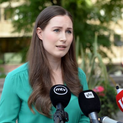 En kvinna med axellångt mörkt hår i en grön blus ser allvarlig ut och pratar mot utsträckta mikrofoner med olika mediebolags logotyper på sig.