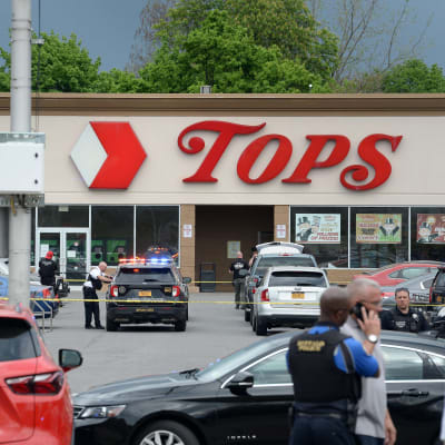 En parkeringsplats utanför en affär som heter Tops har spärrats av, en polis lutar mot en polisbil.