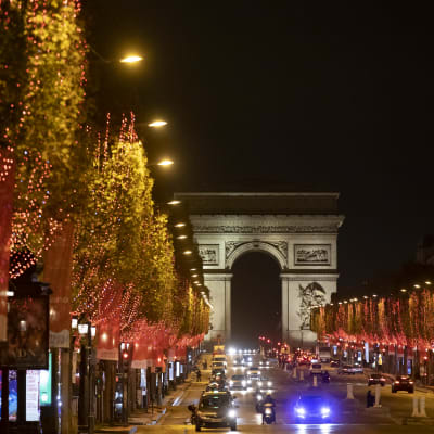 Bild på Triumfbågen i Paris. På Champs-Elysées kör bilar. Gatan omges av julljus i träden.