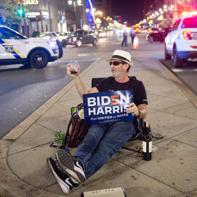 Enman sitter på en refuge med ett vinglas i handen. I andra handen håller han en skylt med texten "Biden Harris".