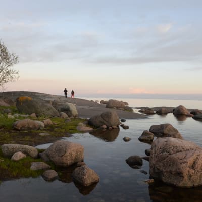 Kaksi ihmistä seisoo selin kivisellä luodolla. Etualalla on rantakiviä. On hieman hämärää ja taivaanranta on punertava.