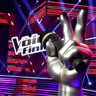 Symbolen för The Voice of Finland i förgrunden, i bakgrunden scenen som används i programmet.