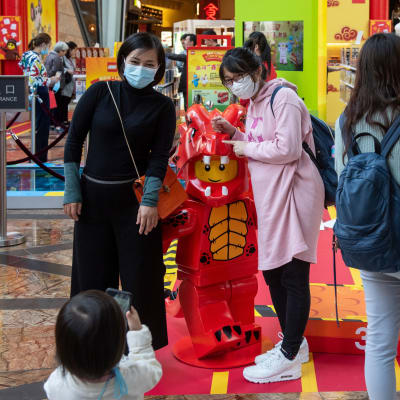 Två kvinnor med munskydd poserar för ett litet barn som tar bild. Kvinnorna står kring en röd legofigur. 